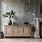 image of limed oak sideboard with two doors on either side and three drawers in the middle, with faux plants on top, pushed against a distressed grey-blue wall