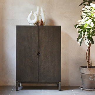 image of dark wood cabinet in front of a beige wall, topped with decorative accessories, next to a tall plant with light shining onto it