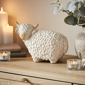 large ceramic neautral coloured highland cow displayed on a sideboard, surrounded by cosy candles and a potted plant.