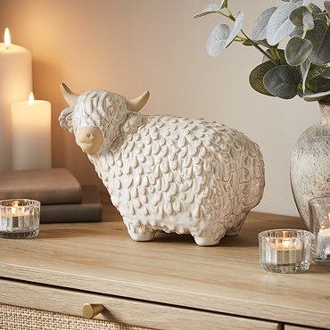 trio of neutral coloured highland cows in three different sizes, displayed on a sideboard surrounded by candles and plants.