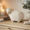 large ceramic neautral coloured highland cow displayed on a sideboard, surrounded by cosy candles and a potted plant.