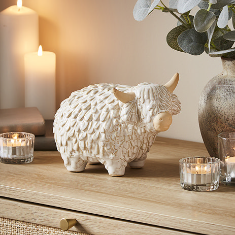 medium natural coloured ceramic highland cow displayed on a sideboard, surrounded by lit candles.