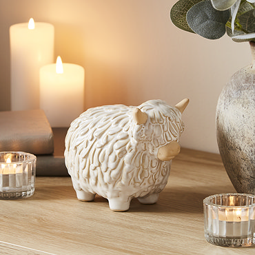 trio of ceramic highland cows in three sizes on a warm wood sideboard, surrounded by cosy candles.