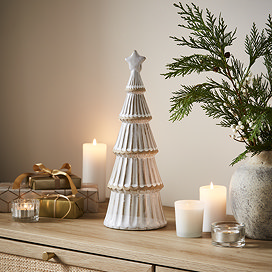 large ceramic christmas tree in a matte finish with a star on top, displayed on a sideboard surrounded by candles, small presents and a potted plant.