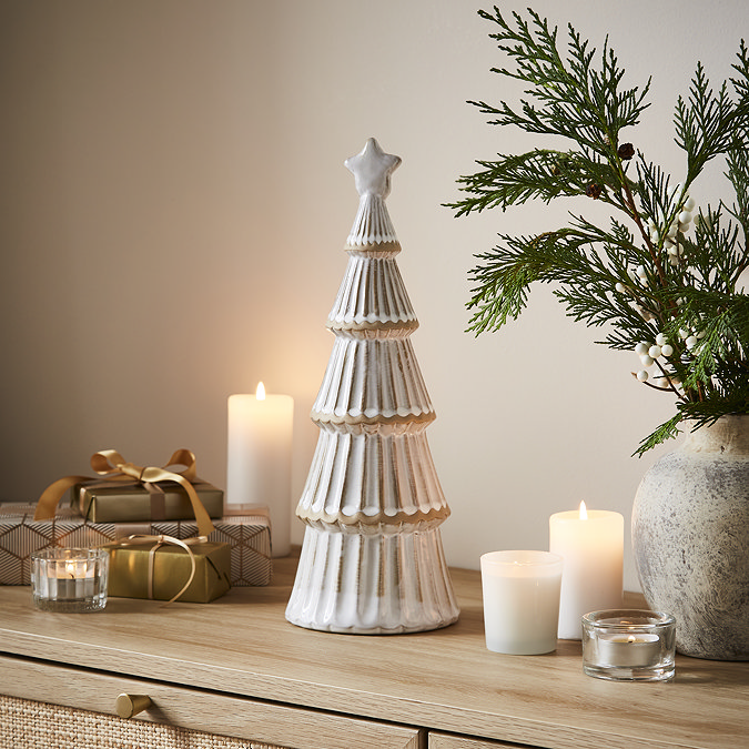 large ceramic christmas tree in a matte finish with a star on top, displayed on a sideboard surrounded by candles, small presents and a potted plant.
