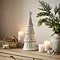 large ceramic christmas tree in a matte finish with a star on top, displayed on a sideboard surrounded by candles, small presents and a potted plant.