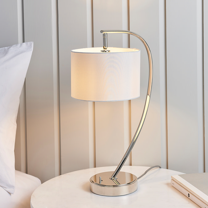image of gently arched nickel table lamp with faux silk white shade on a cream bedside table in front of a cream panelled wall