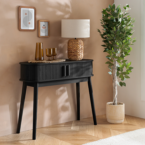 black console table with fluted drawers and thin legs, topped with lamp and glasses, next to a tall plant and pushed up against a light brown wall