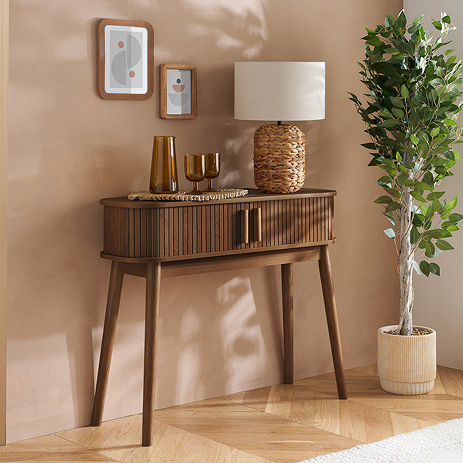 walnut console table with fluted sides and two doors, topped with lamp and glasses, up against a light brown wall decorated with frames and a large potted plant next to it