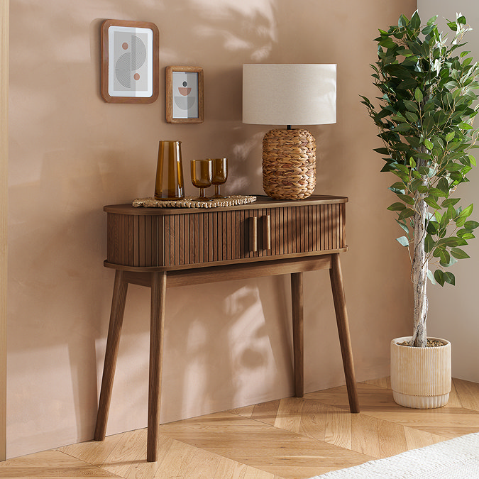 walnut console table with fluted sides and two doors, topped with lamp and glasses, up against a light brown wall decorated with frames and a large potted plant next to it