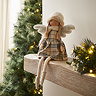 side on photo of decorative angel in cream tartan dress sitting on mantelpiece with christmas tree out of focus in the background