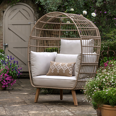 image of rounded wicker egg chair in garden with plush cream cushions and acacia wood legs