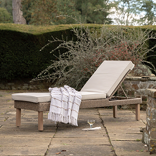 image of outdoor sun lounger, with rattan base and plush cream cushion, draped with throw