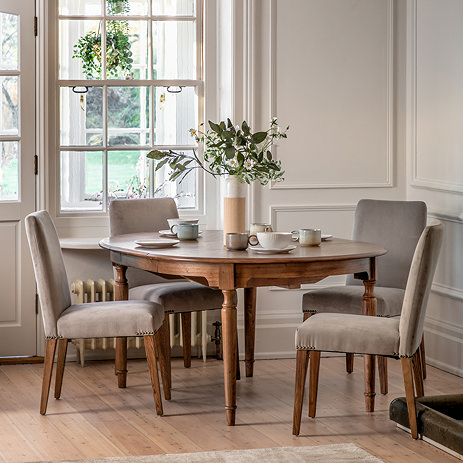 image of round mindi wood dining table with upholstered grey velvet dining chairs around it, on wood flooring of dinign room