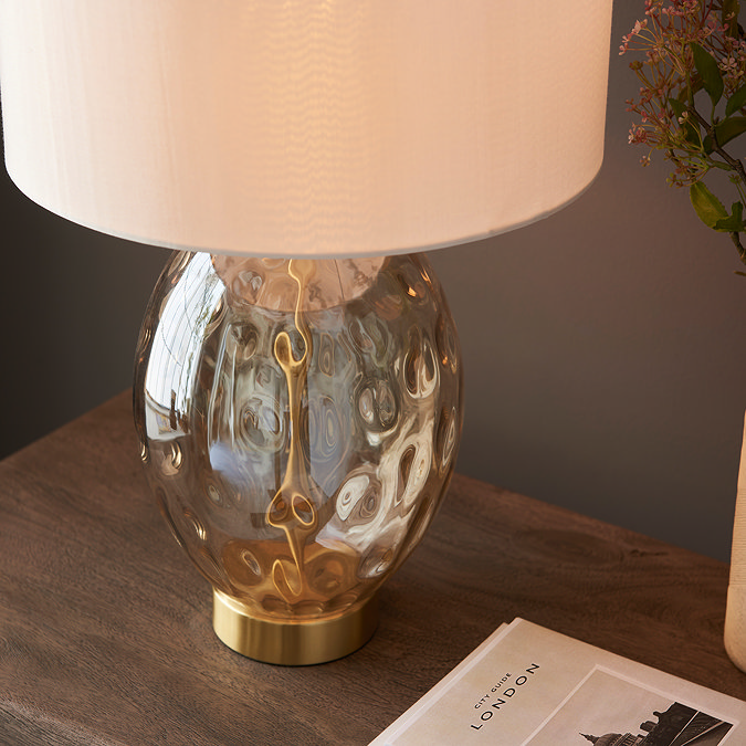 close up image on dimpled champagne gold body of table lamp, warmly lit by bulb inside, on a warm wooden side table