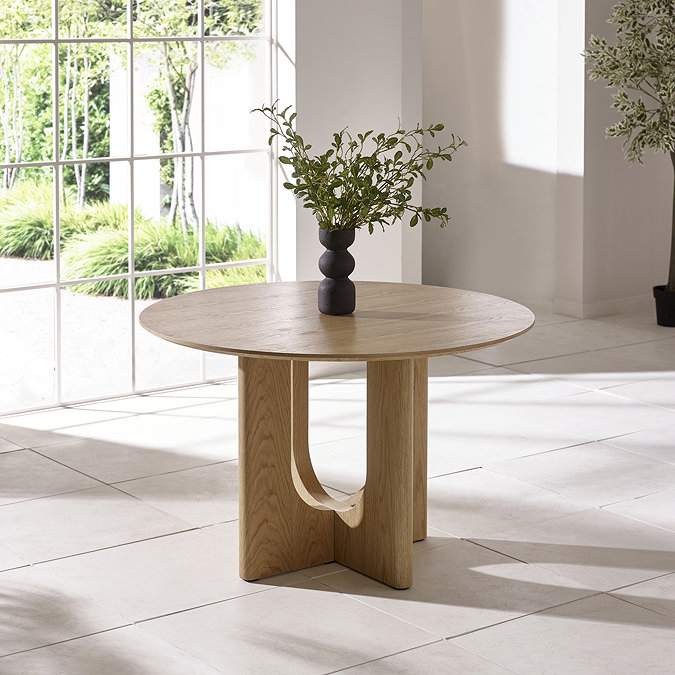 image of round oak dining table without chairs around it, on cream marble flooring with modern black vase on top