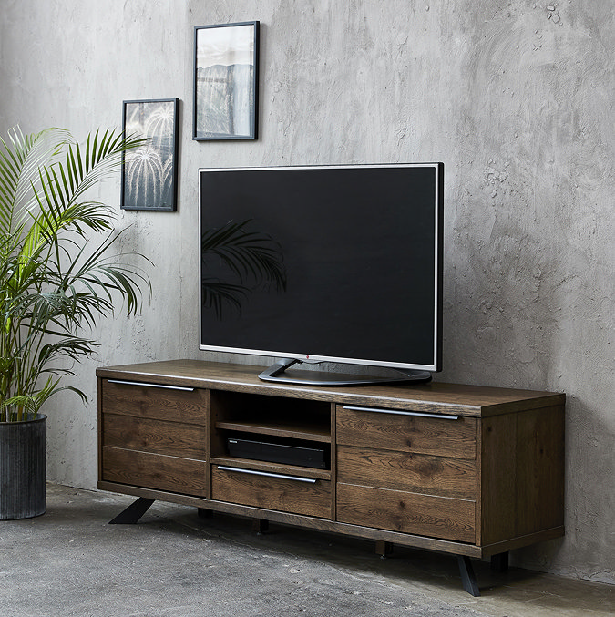 image of dark oak panelled tv unit with tv on top, two drawers on either side of a middle shelf and drawer, pushed against a grey wall adorned with picture frames and a tall plant in the left corner