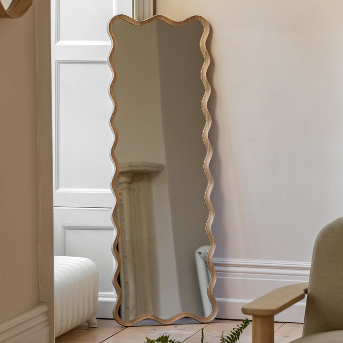 image of light oak tall leaner mirror with wavy silhouette, against a cream hallway wall