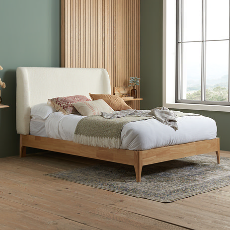side on angled image of large double wooden bedframe with white boucle headboard and mattress dressed up on top.