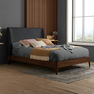 side on image of dark wood bedframe with grey boucle curved headboard, dressed with mattress and dark sheet and warm accent cushions