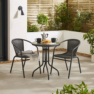 image of black steel bistro set with wicker backed chairs, on stone patio surrounded by plants