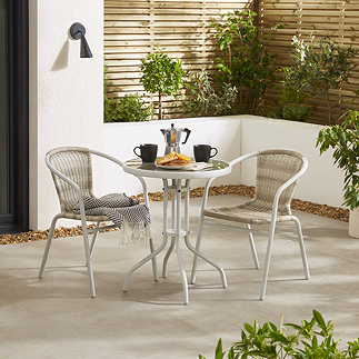 image of white steel bistro set with wicker seating, on a stone patio surrounded by hedges