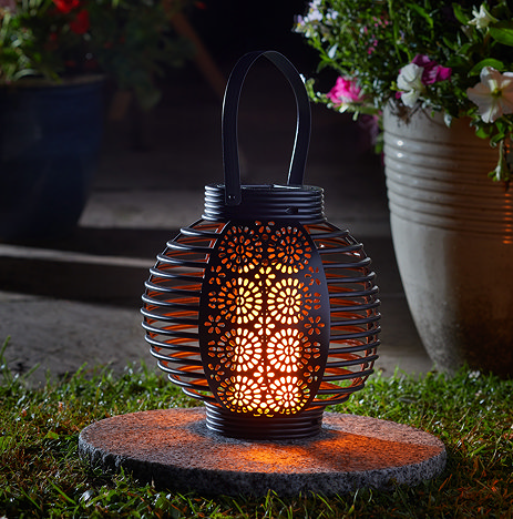 image taken at night of round black garden lantern with intricate floral pattern cut out, with amber light glowing through