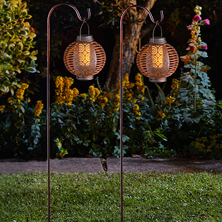 image of two bronze coloured lanterns on shepherd's hooks in garden at night, emitting amber LED light