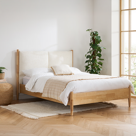 image of oak bedframe with linen headboard, against cream wall next to potted plant in bedroom