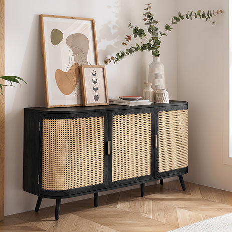 image of three door black sideboard with rattan front, against a pale taupe living room wall with abstract art and faux plants on top