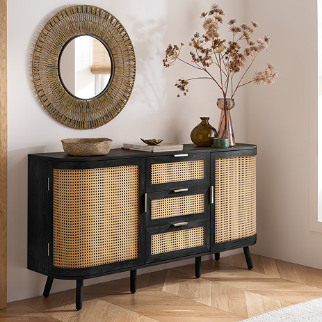image of black rattan sideboard with 2 doors, one either side of three middle drawers, pushed up against a light taupe wall with mirror hanging above it and decorative vases on top