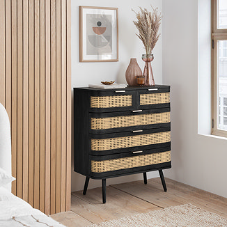 image of 5 drawer black chest with rattan front and gold handles, against a light grey bedroom wall with decorative vases on top