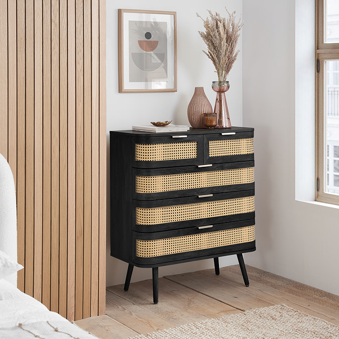 image of 5 drawer black chest with rattan front and gold handles, against a light grey bedroom wall with decorative vases on top