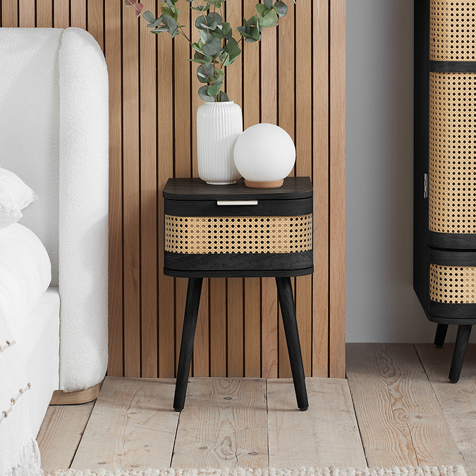 image of black bedside table with single rattan-fronted drawer on a wooden floor and topped with globe light and medium white vase
