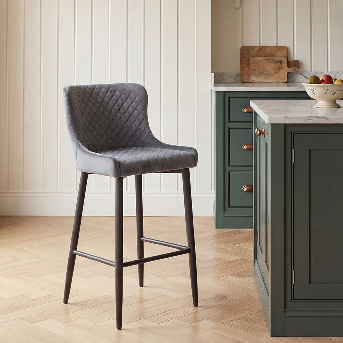 image of single grey velvet bar stool with black legs and harlequin stitching, next to modern country counter on herringbone light wood flooring