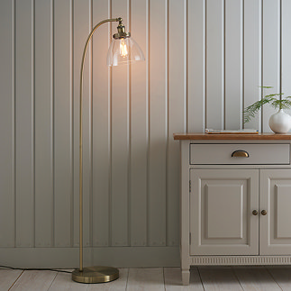 image of gently arched antique brass floor lamp with clear shade, stood in front of grey panelled wall next to grey sideboard, diffusing warm light