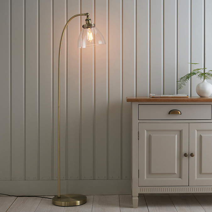 image of gently arched antique brass floor lamp with clear shade, stood in front of grey panelled wall next to grey sideboard, diffusing warm light