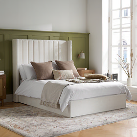 image of ottoman bed with tall cream linen headboard, covered in natural coloured cushions and throws