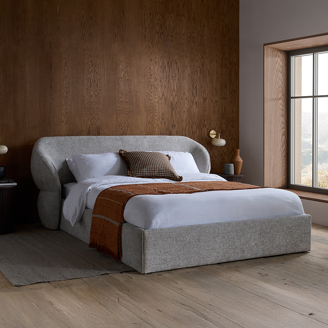 image of grey fabric bed with rounded headboard, against dark brown wall