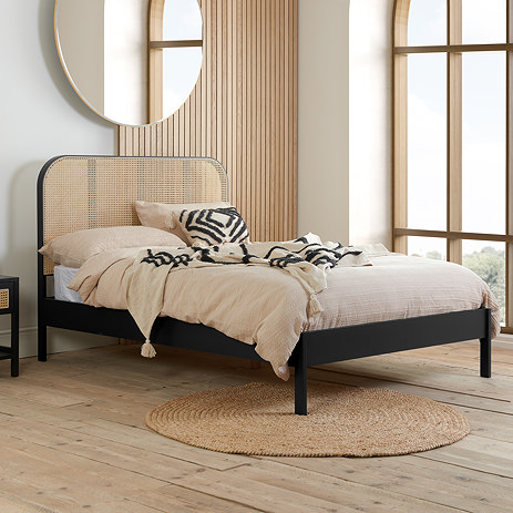 side on image of black rattan double bed with cream duvet on and black patterned cushions