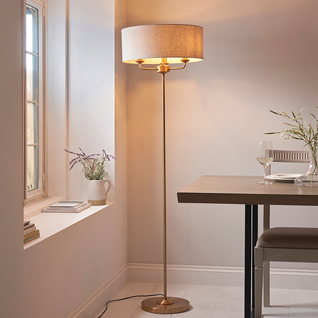 image of vintage brass floor lamp with linen coloured shade over 3 arms, emitting a warm glow at the end of a dark oak dining table