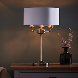 image of antique brass 3 arm table lamp on a bedside table, emitting a warm low against a dark painted wall