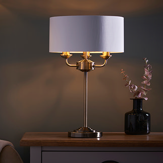 image of antique brass 3 arm table lamp on a bedside table, emitting a warm low against a dark painted wall