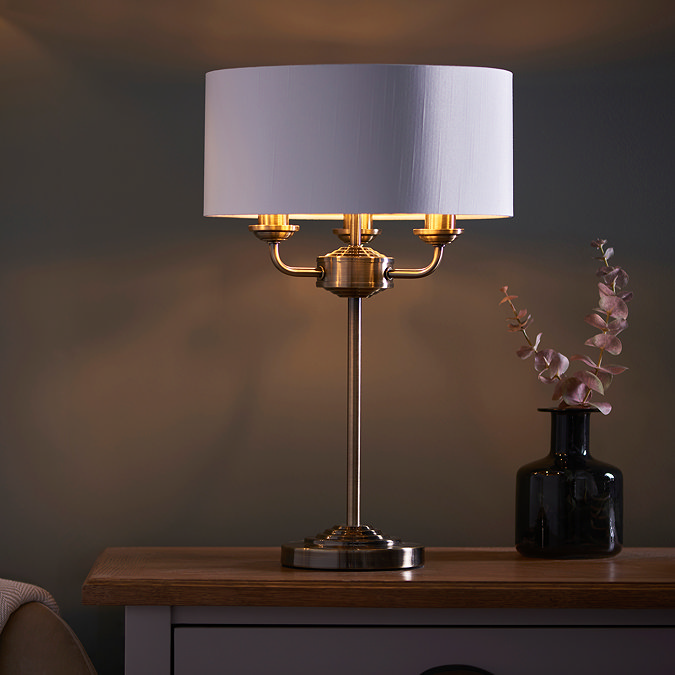 image of antique brass 3 arm table lamp on a bedside table, emitting a warm low against a dark painted wall