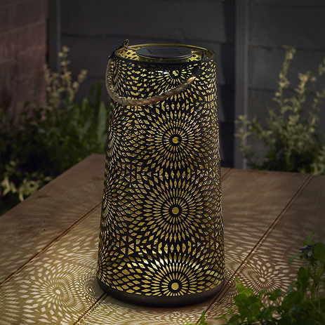image of tall lantern with swirl floral patterns cut out and warm white light shining out to create patterns on wooden outdoor table