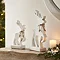 white wooden standing reindeer next to larger version of same product, stood on a mantlepiece with candles and garlands.