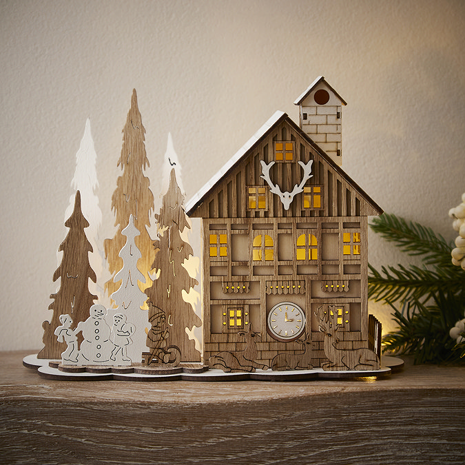 front on image of wooden house decoration on a mantlepiece with deer outside the front door, trees, and children playing with a snowman. lit up from the inside by warm white LEDs.