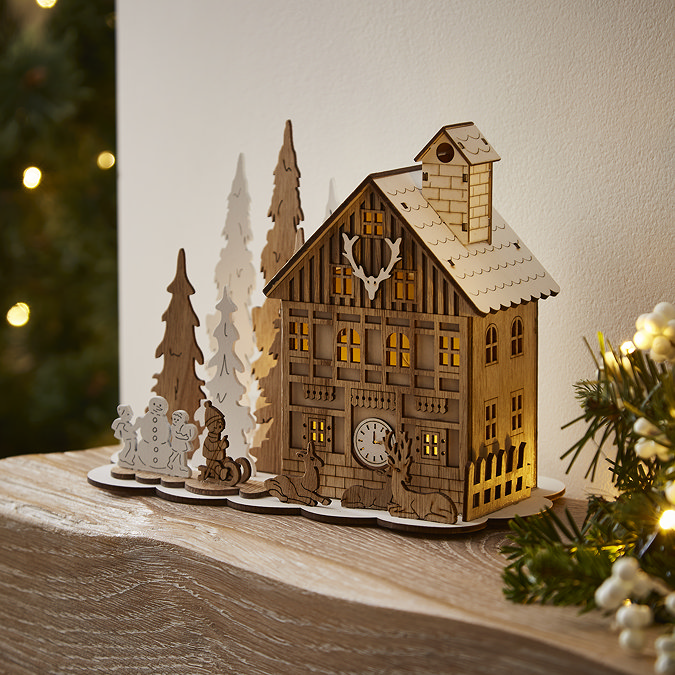 slightly angled photo of wooden house decoration stood on a mantlepiece, next to wooden trees and wooden children figures decorating a snowman