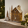 slightly angled photo of wooden house decoration stood on a mantlepiece, next to wooden trees and wooden children figures decorating a snowman