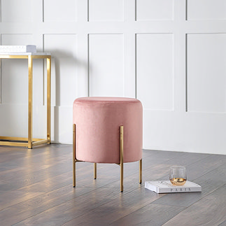 image of pink velvet pouffe with gold accent legs, stood on warm wooden flooring with gold console table in the background